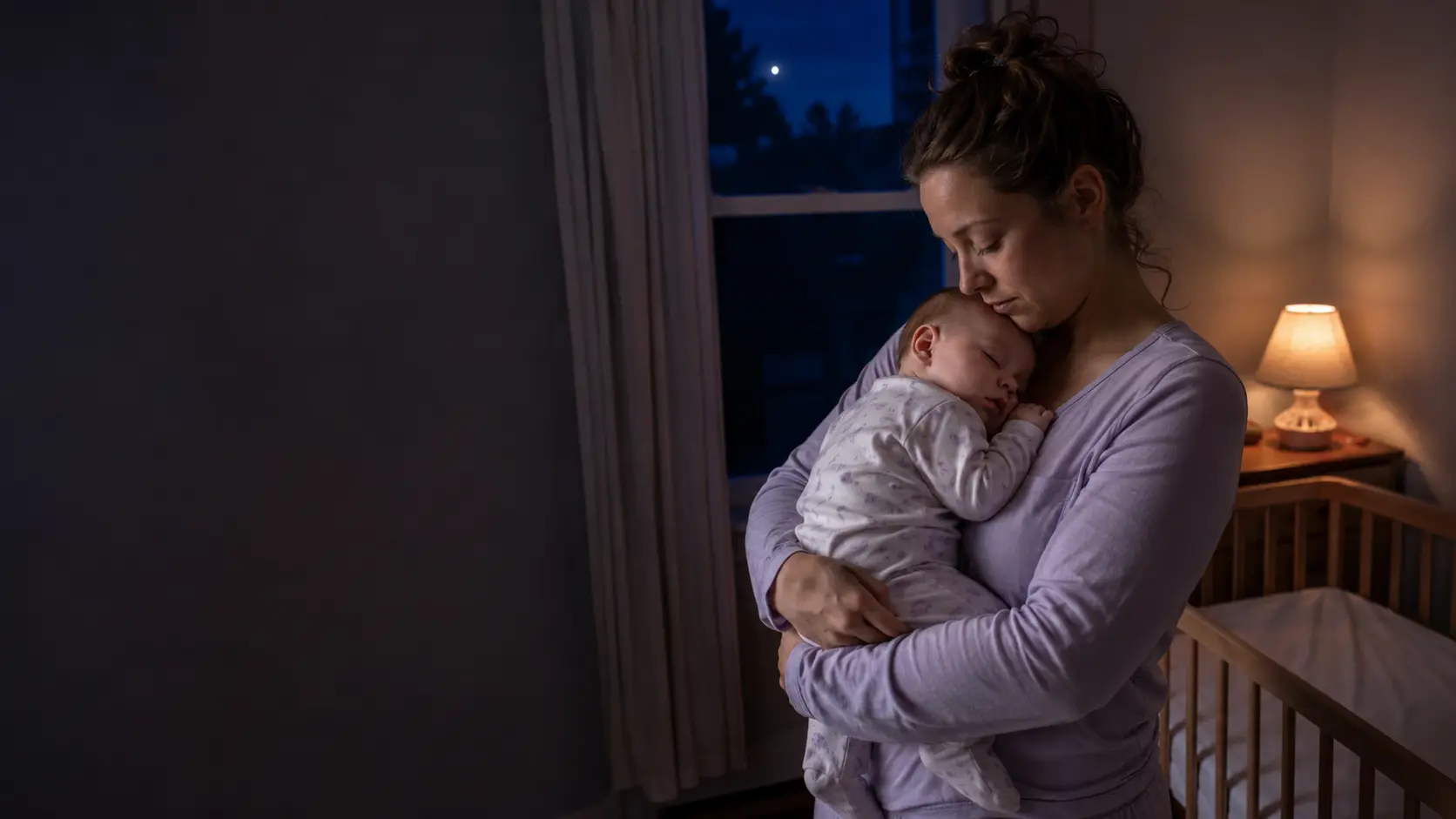 A tired, attentive mother holding her sleeping baby beside a bare crib in a moonlit nursery