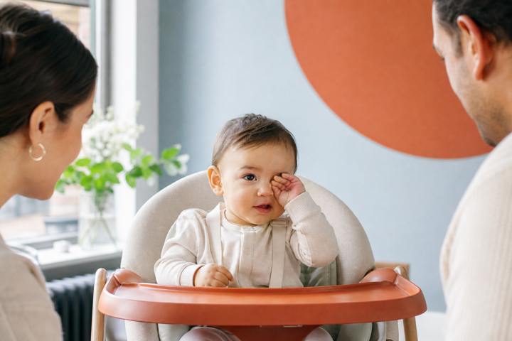 Baby rubs one eye in a high chair while two caregivers watch