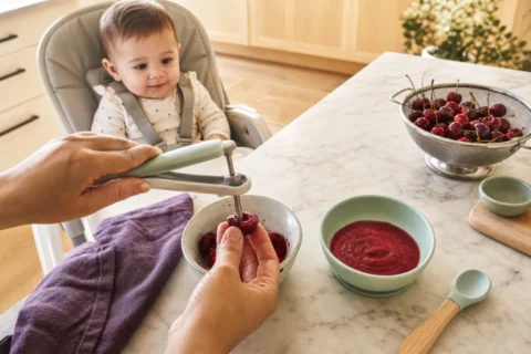 Caregiver pits a cherry beside a bowl of mashed cherries while a baby watches from a high chair.