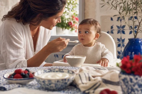 Baby tastes a small spoonful of yogurt in a high chair while a parent watches for the baby's reaction.
