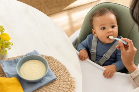 Caregiver spoon-feeds smooth grits to a baby seated upright in a high chair.