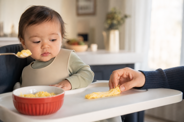 Baby turns away as a caregiver offers scrambled egg at a kitchen high chair.