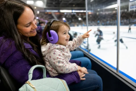 A toddler in purple hearing protectors points toward the ice while sitting with a caregiver at a hockey game.