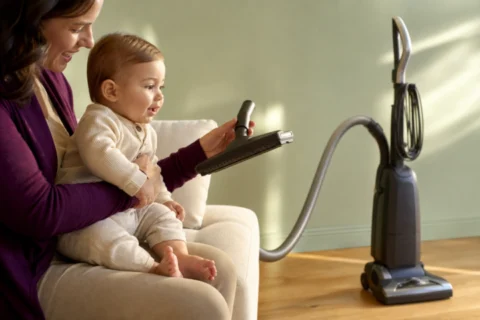 Caregiver securely holds an awake baby who watches an unplugged vacuum and its floor attachment.