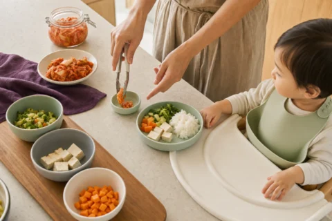 Caregiver portions kimchi into a small tasting bowl beside a seated toddler's rice, tofu, and vegetables.