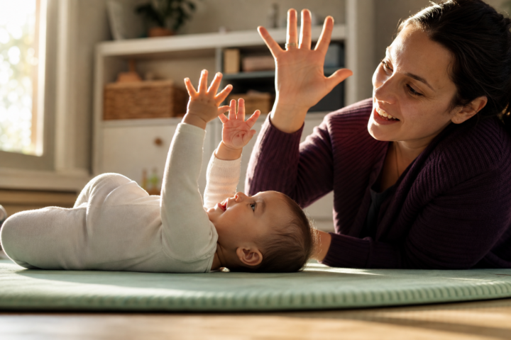Awake baby studies both hands while a parent mirrors the gesture beside a mint play mat.