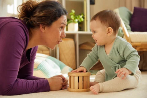 Baby makes a playful growling face while a caregiver mirrors the sound during floor play.