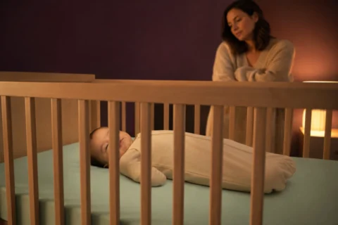 Caregiver watches a baby sleeping on their back with one eyelid slightly open in an empty crib.