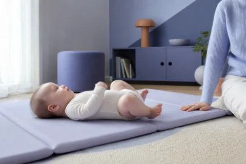 Awake baby rests with bent legs on a lavender play mat beside a parent