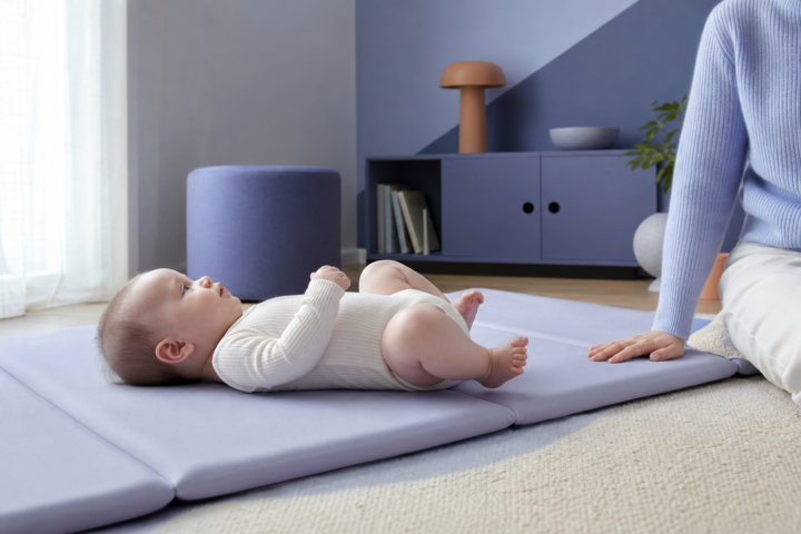 Awake baby rests with bent legs on a lavender play mat beside a parent