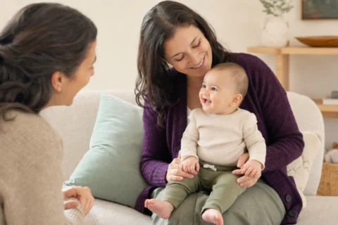 Awake baby smiles toward a visiting family member while seated safely on a parent's lap.