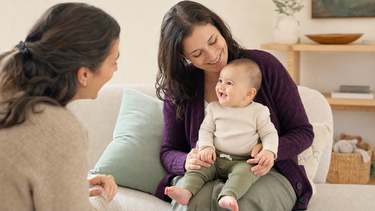 Awake baby smiles toward a visiting family member while seated safely on a parent's lap.