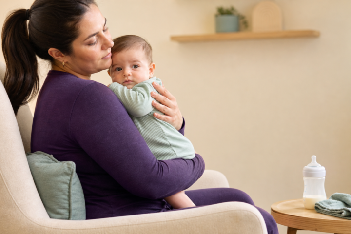 Caregiver holds an awake baby upright beside a bottle and burp cloth in a calm nursery.