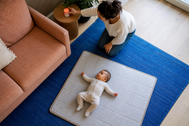 A parent adjusts a red night-light beside an awake baby lying on a floor mat in a bright family room.
