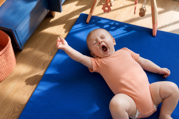 Four-month-old baby yawns and stretches on a cobalt play mat in a sunlit family room.