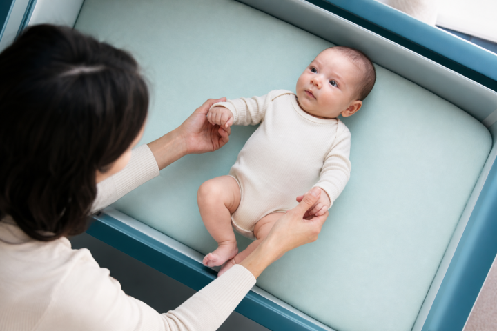 Parent holds an awake young baby's hands in a bright aqua bassinet