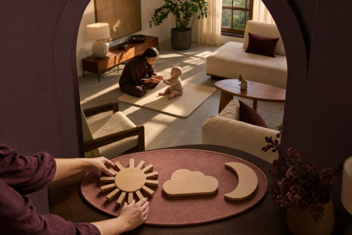 Adult hands arrange wooden sun, cloud, and moon symbols while a mother supervises her seated baby in a sunlit living room.