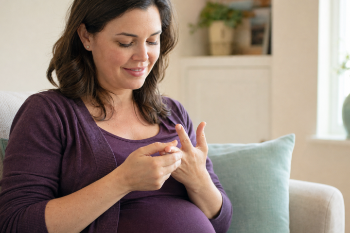 Pregnant woman in a deep purple top gently massages a finger while seated in a cream room with a mint pillow.