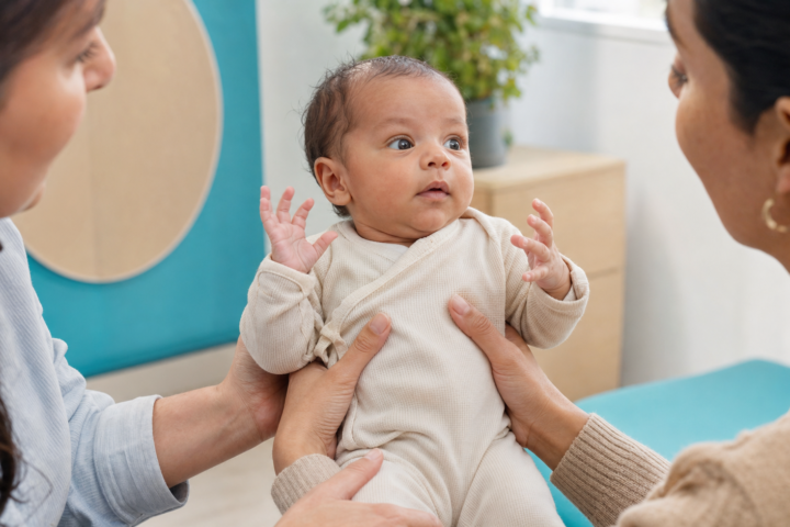 Awake baby lifts both hands while two caregivers provide support in a bright turquoise-accented coaching room.