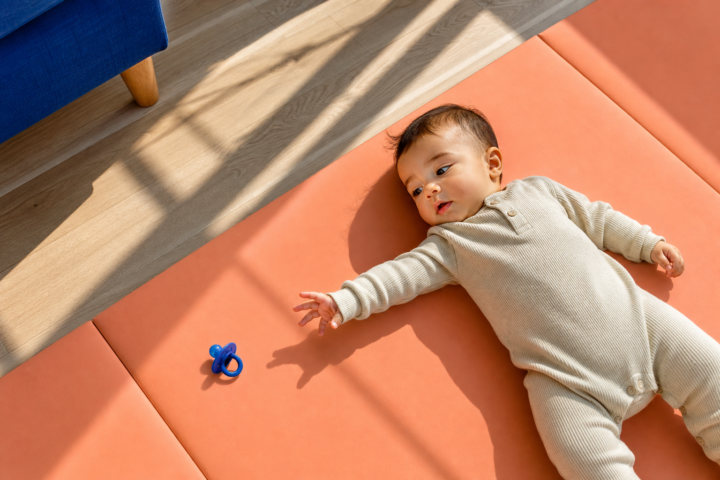 Awake baby reaches toward a blue pacifier on a peach play mat in a sunlit family room