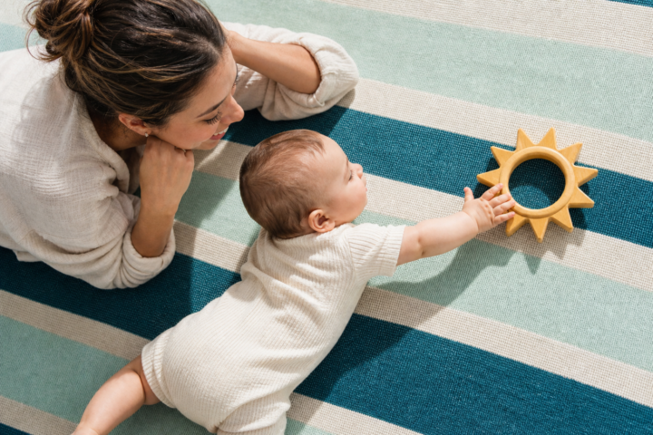 A six-month-old baby reaches for a wooden sun ring during supervised floor play beside a parent.