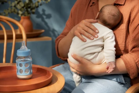 Sleeping infant rests upright against a caregiver beside a bottle in warm morning light.