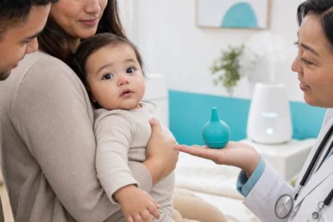 Two caregivers hold their baby as a pediatrician displays a turquoise nasal bulb in a bright clinic