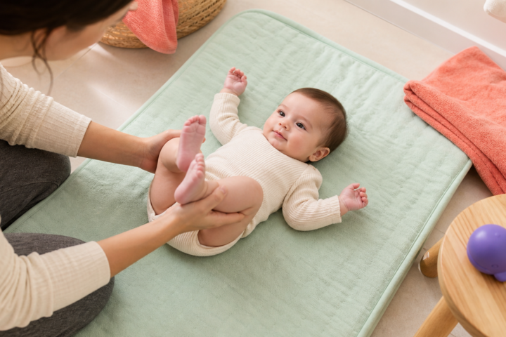 Caregiver gently bicycles an awake baby's legs on a padded mat after bath time.