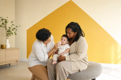 Two caregivers comfort an alert baby with visible red spots and a cool washcloth in a bright ochre room.