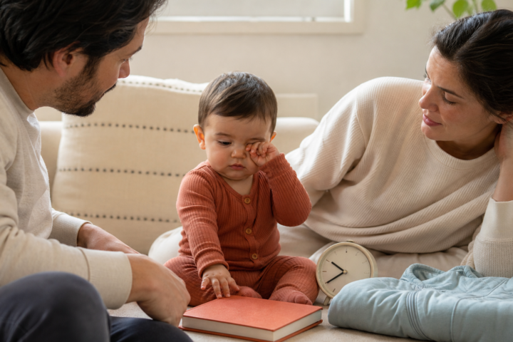 Drowsy baby rubbing one eye between two caregivers beside a clock, bedtime book, and folded sleep sack