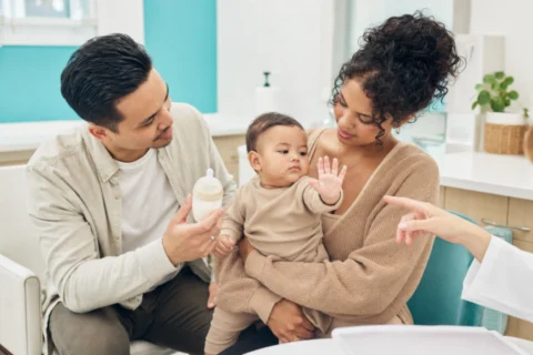 Baby turns from a bottle and raises one hand while seated between two caregivers.