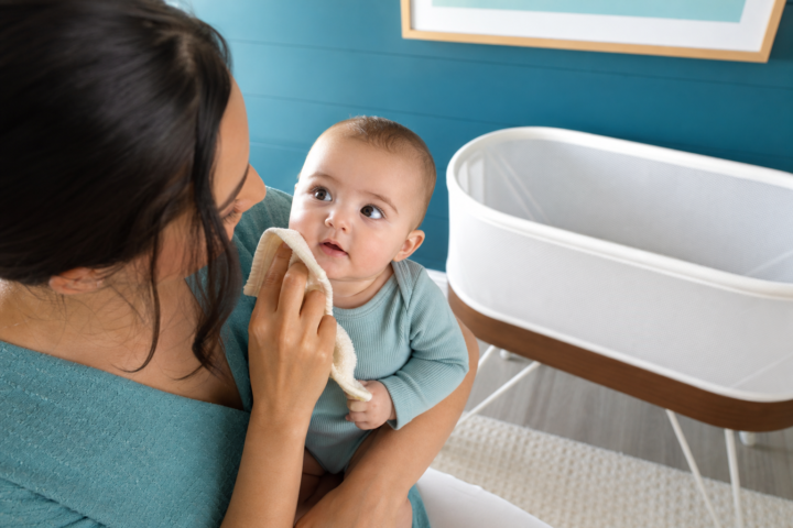 Parent gently wipes an alert baby's cheek beside an empty bare bassinet in a bright teal nursery.