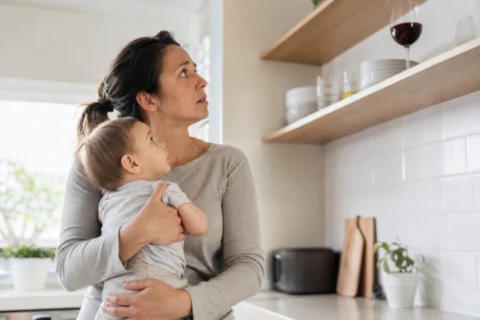 Parent holding a baby in a kitchen while a wine glass sits high on a shelf in the background.