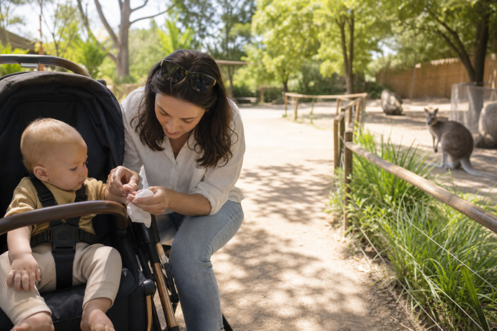 Caregiver checking a baby's hand beside a stroller on a zoo walkway with a kangaroo in the background.
