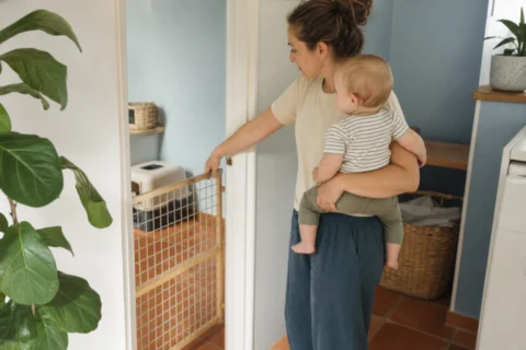 Caregiver holding a baby near a pet gate and a closed litter box area in a bright hallway.