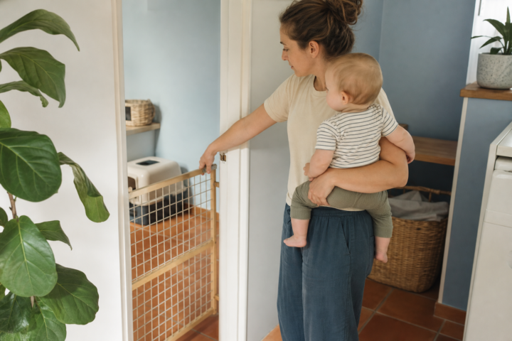 Caregiver holding a baby near a pet gate and a closed litter box area in a bright hallway.