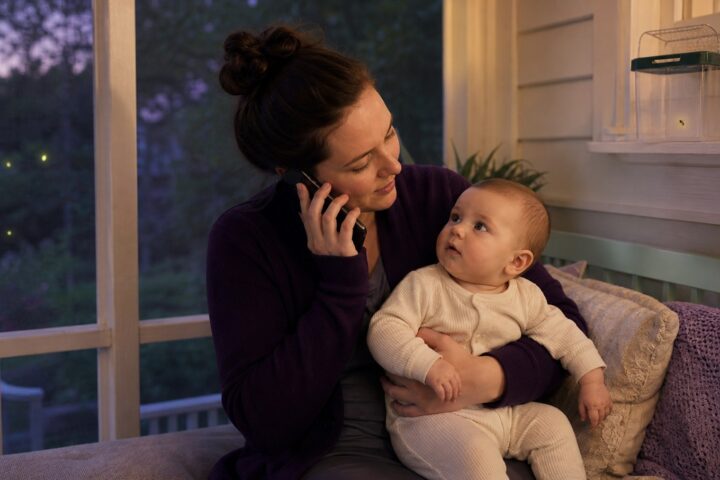 Mother holding her awake baby while calling for advice on a screened porch with a firefly secured in a high container