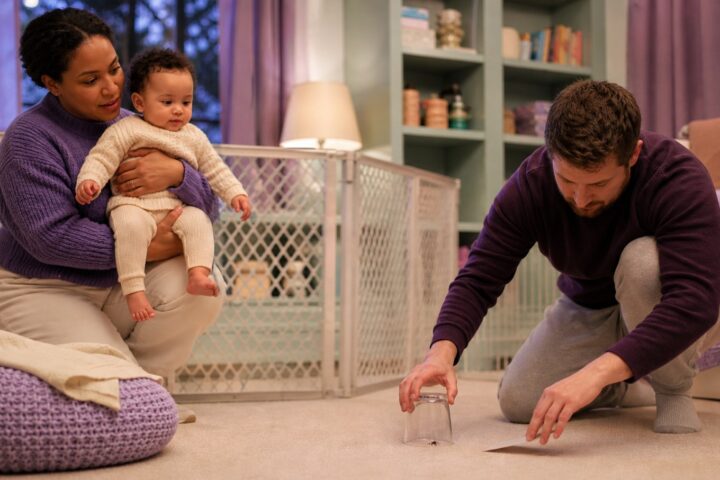 Mother carrying her awake baby toward a safety gate while a father contains a small spider under a clear cup