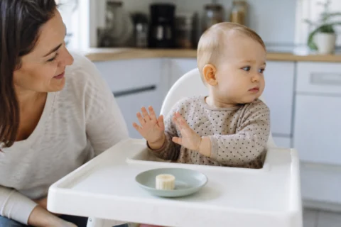 Baby sitting in a high chair and turning away from a small banana slice on a plate.