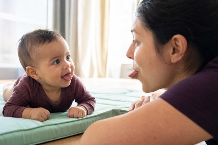 Baby doing supervised tummy time and sticking out his tongue while his mother copies the expression.
