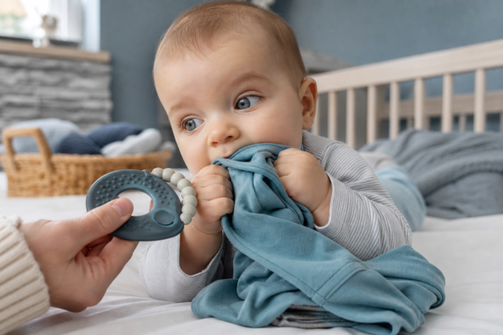 A baby chews on a blue shirt while a parent offers a blue teether in a nursery.