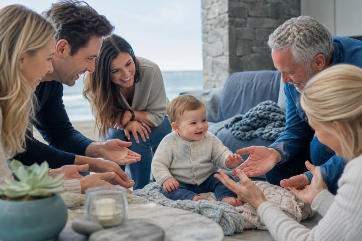 A smiling baby sits on a blanket surrounded by friendly adults reaching in from around the frame.
