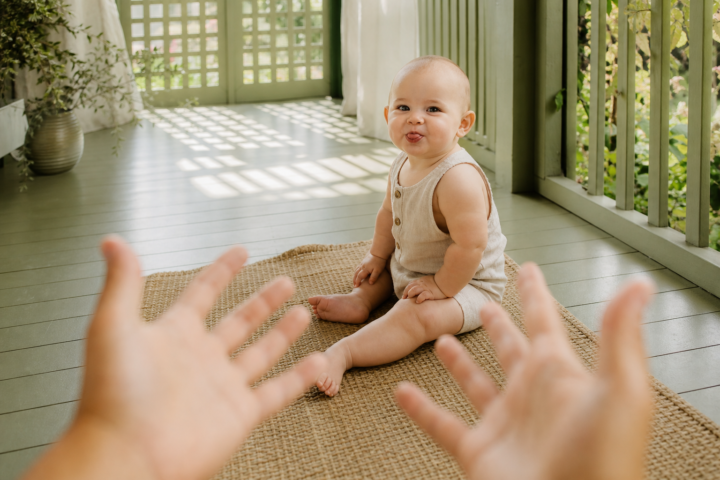 Baby blowing a raspberry toward caregiver hands on a sage porch.