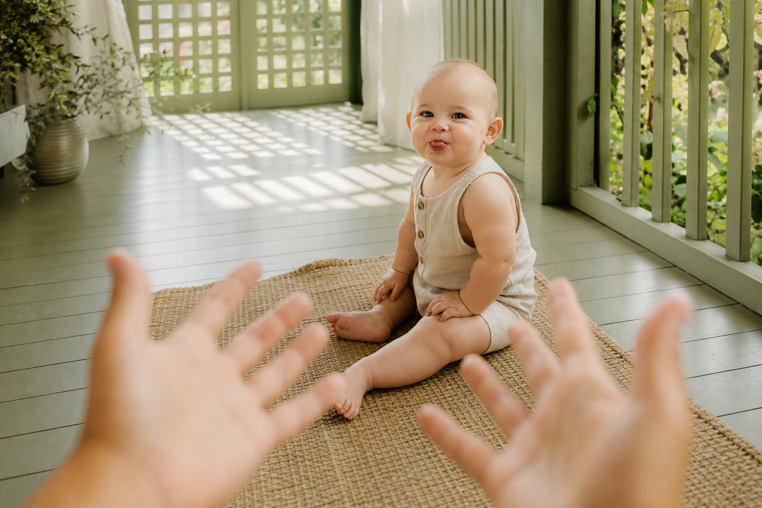 Baby blowing a raspberry toward caregiver hands on a sage porch.
