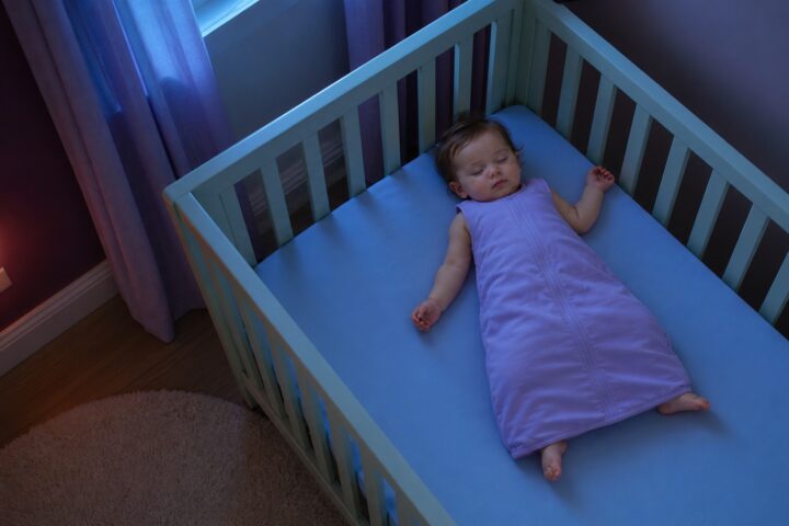 A baby in a lavender sleep sack lies on their back near the corner of an otherwise empty crib.