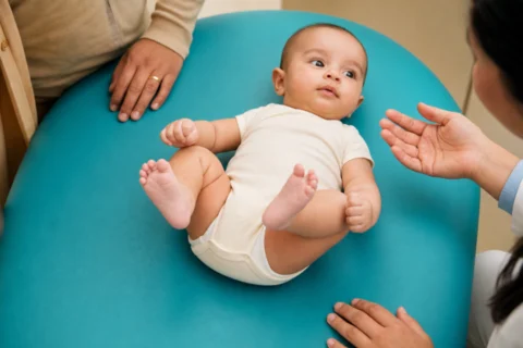 Awake baby lifts both bent legs on a turquoise pediatric exam pad while caregivers stay nearby