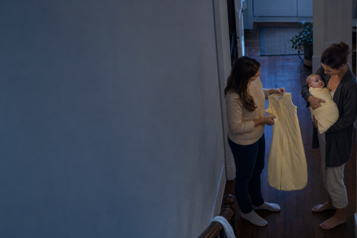 Sleep coach shows a pale yellow sleep sack to a caregiver holding a swaddled baby in a blue-hour hallway.
