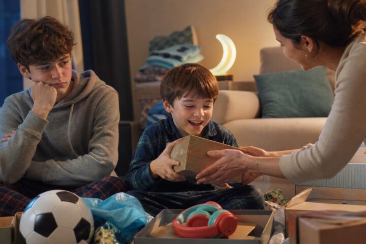 Mother hands another present to a delighted younger brother while his older sibling watches empty-handed.