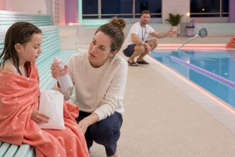 Queasy child in a coral towel holding a sickness bag while a parent offers water beside a cleared swimming pool