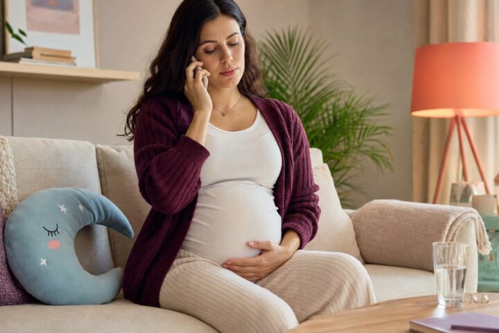 Pregnant woman sitting on a sofa with one hand on her lower abdomen while speaking on the phone.